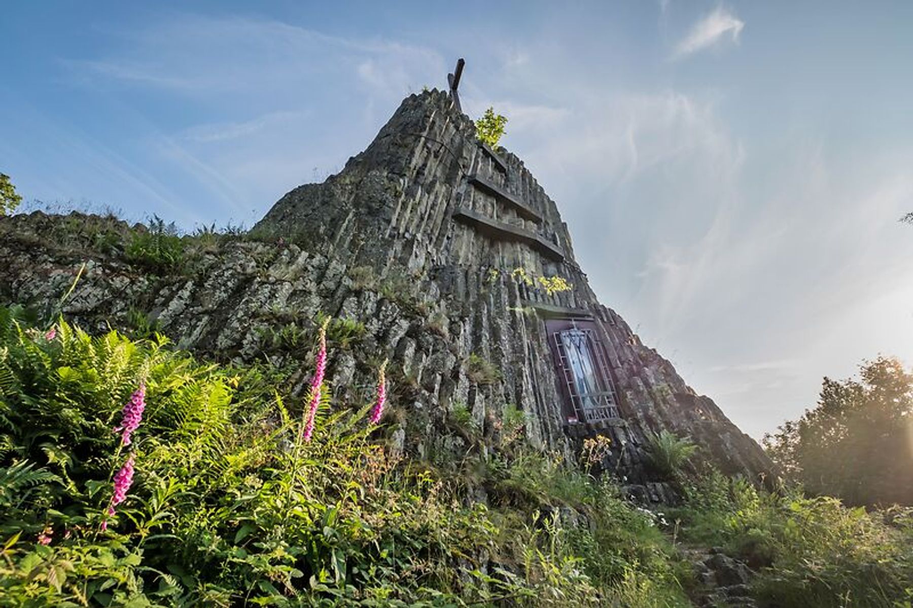 A rocky hill with a metal door built into its side, surrounded by green plants and purple wildflowers, under a blue sky with sunlight streaming in from the right.